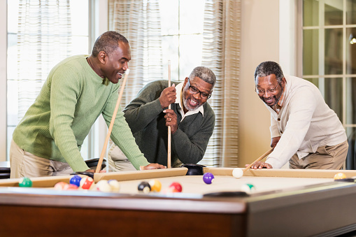 Three mature and senior African-American men in their 50s and 60s having fun shooting pool. One man is about to hit the cue ball with his cue stick while his friends smile and watch.