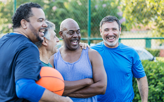 A multi-ethnic group of middle-aged and senior men on an outdoor basketball court. The Hispanic man looking at the camera is in his 40s. The African-American man standing beside him with his arms crossed is in his 50s.