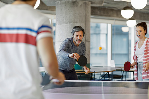 Mature businessman playing table tennis with colleagues. Male and female coworkers are having fun at workplace. They are in creative office.