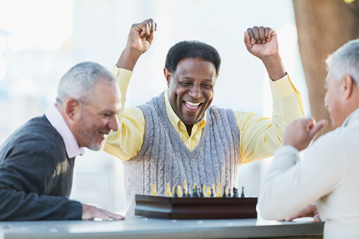 A group of three middle-aged and senior multi-ethnic men in their 50s and 60s playing a game of chess. The view if from over the shoulder of the senior Hispanic man. His African-American opponent is laughing, fists in the air, declaring victory.