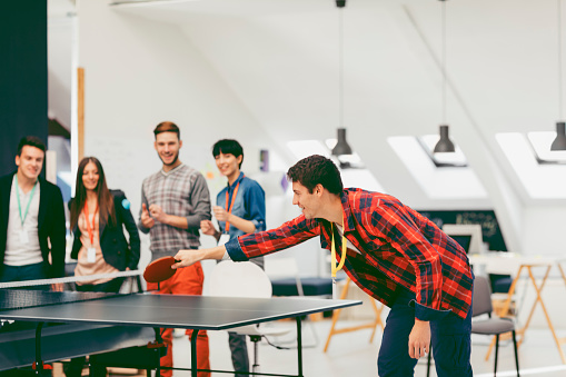 Group of young business people running new start up company. They have table tennis in their office. Two colleague playing table tennis and other coworkers cheering. Standing on one side of the table and smiling. Focus on male hitting the ball.