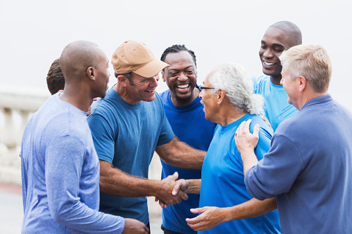 Multi-ethnic group of men of mixed ages wearing casual, blue shirts, outdoors.  Everyone is congratulating the senior Hispanic man (in his late 70s) wearing eyeglasses, shaking his hand and patting him on the back.